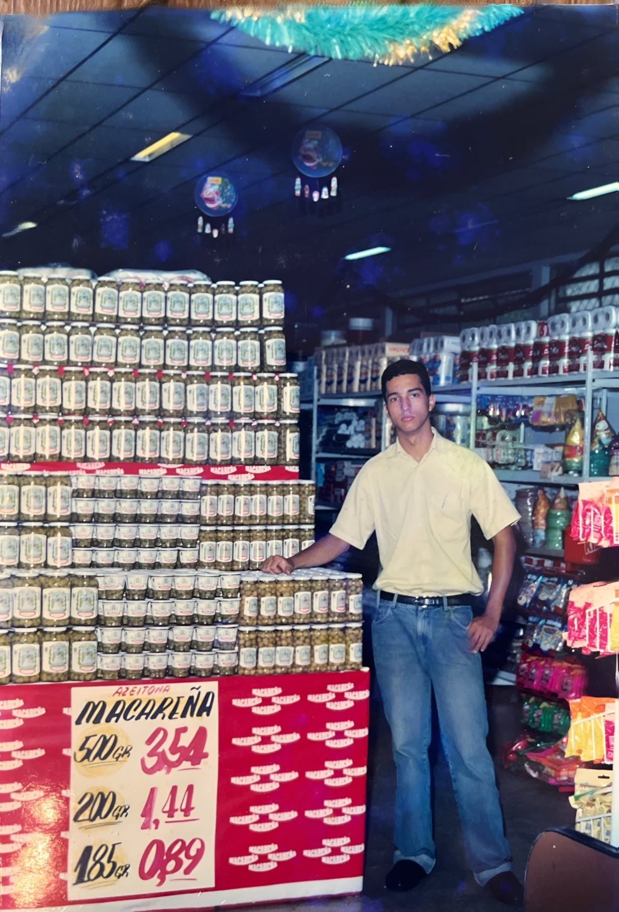 Jonathas jovem de camisa amarela ao lado de uma exposição de azeitonas em um supermercado